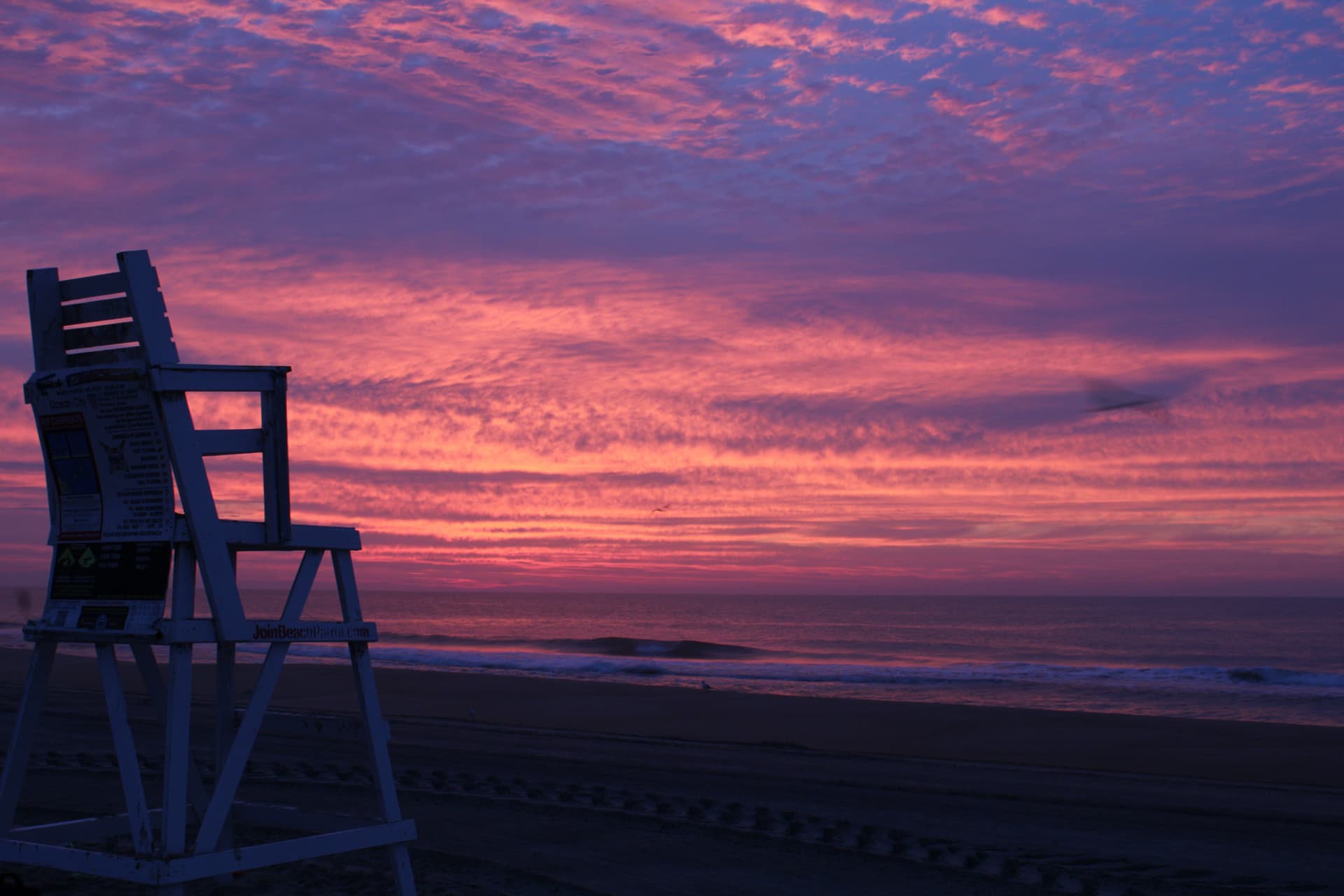 A purple and pink sunrise lighting up the sky with a silhouette of a life guard chair in OC, MD.